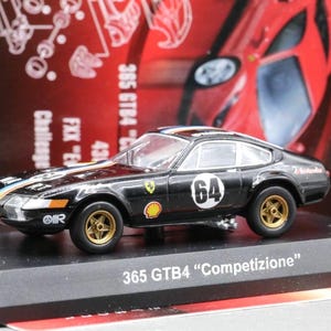 May include: A black Ferrari 365 GTB4 "Competizione" model car with gold wheels and the number 64 on the side. The model sits on a black display stand. The car has racing stripes.