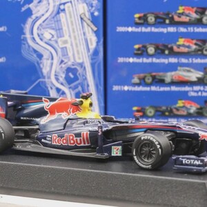 May include: A detailed model of a Red Bull Formula 1 race car. The car is primarily dark blue with red and yellow accents, featuring the Red Bull logo. It sits on a black display stand, with visible sponsor logos and tire details.