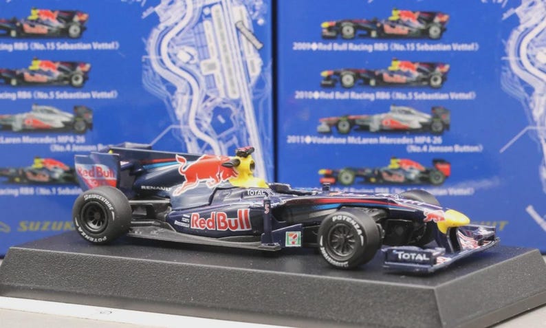 May include: A detailed model of a Red Bull Formula 1 race car. The car is primarily dark blue with red and yellow accents, featuring the Red Bull logo. It sits on a black display stand, with visible sponsor logos and tire details.