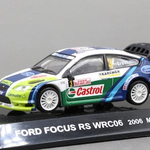 May include: A die-cast Ford Focus RS WRC06 rally car model in white, blue, and lime green livery. The car features Castrol, Karcher, and Ford branding, the number 3, and a rear spoiler. It is mounted on a black display base labelled FORD FOCUS RS WRC06 2006 Monte carlo.