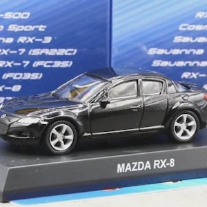 Może przedstawiać: Czarny model samochodu Mazda RX-8 ze srebrnymi kołami i szczegółowym nadwoziem, umieszczony na czarnej podstawie ekspozycyjnej z białym napisem MAZDA RX-8. W tle znajduje się niebieskie pudełko z listą różnych nazw modeli samochodów marki Mazda.