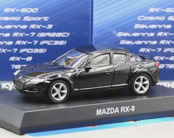 Puede incluir: Un coche a escala Mazda RX-8 de color negro con ruedas plateadas y un exterior detallado, montado sobre una base de exposición negra con el texto MAZDA RX-8 impreso en blanco. El fondo muestra una caja azul con una lista de varios modelos Mazda.