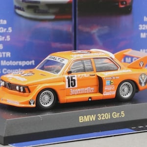 May include: Orange BMW 320i Gr.5 model car with the number 15 on the door. The car has a rear spoiler and sponsor logos. It sits on a black display stand. The background shows other model car boxes.