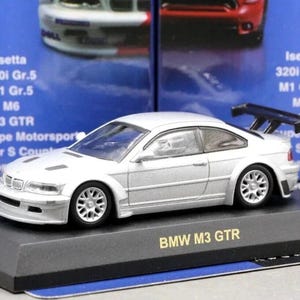 Op de afbeelding: Zilveren BMW M3 GTR modelauto op een zwart displaystandaard. Het model heeft een achterspoiler en gedetailleerde wielen. De standaard is voorzien van het opschrift "BMW M3 GTR". Andere modelautodozen zijn op de achtergrond.