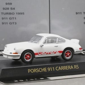 Puede incluir: Un coche a escala Porsche 911 Carrera RS de color blanco con ruedas rojas y una franja lateral roja con la palabra Carrera. El coche está montado sobre una base negra con el texto PORSCHE 911 CARRERA RS impreso en dorado. Fondo de caja gris.