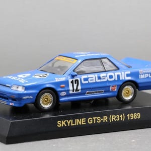 Puede incluir: Un coche de carreras a escala azul Calsonic Impul Nissan Skyline GTS-R (R31) 1989 con el número 12 en el lateral. El coche cuenta con llantas doradas de radios, calcomanías blancas de patrocinadores como Bridgestone e Impul, sobre una base negra.