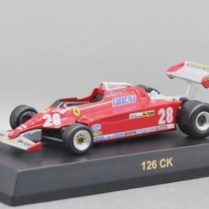Puede incluir: Modelo a escala rojo de un Ferrari 126 CK, con el número 28 en los laterales y la parte delantera. El modelo tiene un alerón blanco y rojo y se exhibe sobre una base negra. La palabra "FIAT" es visible en el coche.