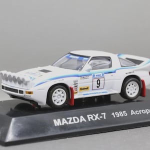 May include: A white Mazda RX-7 model car from 1985, with blue and red accents, and the number 9 on the side. The model car has a rally car design and is mounted on a black base.