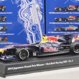 May include: A detailed model of the 2010 Japanese Grand Prix winning Red Bull Racing RB6 No. 5 race car. The car is primarily navy blue with red and yellow accents, and features sponsor logos. It sits on a black display base.