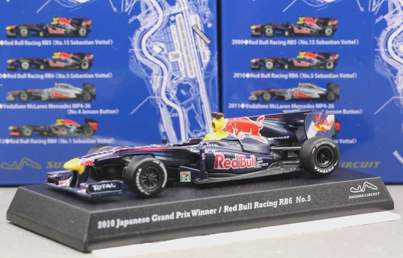 May include: A detailed model of the 2010 Japanese Grand Prix winning Red Bull Racing RB6 No. 5 race car. The car is primarily navy blue with red and yellow accents, and features sponsor logos. It sits on a black display base.