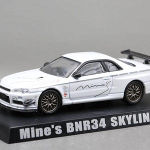 Könnte beinhalten: Ein weißes Modellauto, ein Mine's BNR34 Skyline GT-R, mit dunkelgrauen Vielspeichenfelgen, einem schwarzen Heckspoiler und schwarzen Außenspiegeln. Das Auto ist auf einem schwarzen Sockel mit der Aufschrift Mine's BNR34 SKYLINE GT-R montiert.