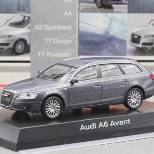 Puede incluir: Un modelo de coche gris Audi A6 Avant sobre un soporte de exhibición negro. El modelo de coche tiene detalles, incluyendo ruedas y ventanas. El texto "Audi A6 Avant" está impreso en el soporte.