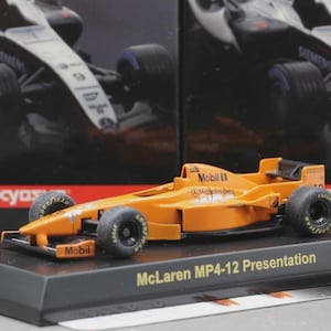 Op de afbeelding: Oranje McLaren MP4-12 Presentation model raceauto op een zwarte displaystandaard. Het model heeft zwarte banden en Mobil-branding. De achtergrond toont een wazige afbeelding van een vergelijkbare auto.