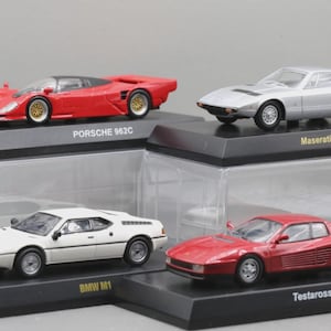 Puede incluir: Cuatro modelos de coches en miniatura en exhibición. Un Porsche 962C rojo, un Maserati Khamsin plateado, un BMW M1 blanco y un Testarossa rojo. Cada coche está sobre un soporte negro con el nombre del coche.