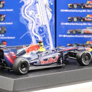 May include: A detailed model of a Red Bull Formula 1 race car, featuring the Red Bull logo and sponsor decals. The car is primarily navy blue with red and white accents, and sits on a black display base.