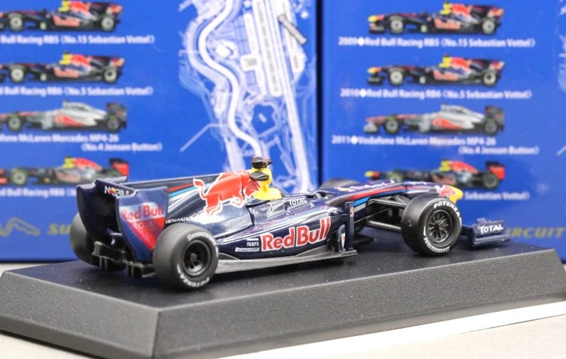 May include: A detailed model of a Red Bull Formula 1 race car, featuring the Red Bull logo and sponsor decals. The car is primarily navy blue with red and white accents, and sits on a black display base.