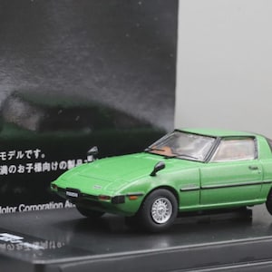 May include: A green diecast model car with silver wheels. The model is a Mazda RX-7, a classic sports car. The model is on a black display stand. The text on the box reads "Mazda Motor Corporation".