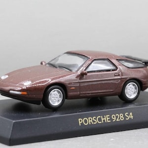 Puede incluir: Un modelo en miniatura de un coche Porsche 928 S4 marrón. El coche de fundición tiene ruedas plateadas y un alerón negro. El modelo se encuentra sobre una base negra con el texto "PORSCHE 928 S4".