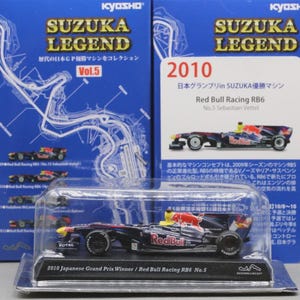 May include: A miniature Red Bull Racing RB6 race car, the 2010 Japanese Grand Prix winner, is displayed in a clear plastic case. The car is primarily navy blue with red and yellow accents, and the text "Red Bull" is visible. The packaging includes the text "SUZUKA LEGEND".