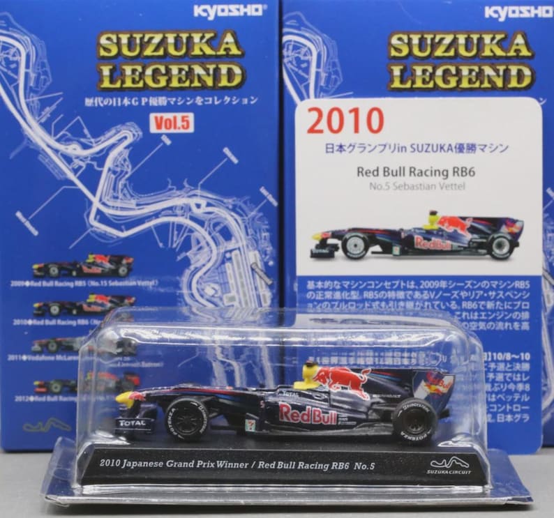 May include: A miniature Red Bull Racing RB6 race car, the 2010 Japanese Grand Prix winner, is displayed in a clear plastic case. The car is primarily navy blue with red and yellow accents, and the text "Red Bull" is visible. The packaging includes the text "SUZUKA LEGEND".