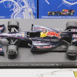 May include: A detailed model of a Red Bull Racing RB6 Formula 1 car. The navy blue car features red and yellow accents, sponsor logos, and the Red Bull logo. The model sits on a black base with text that reads "2010 Japanese Grand Prix Winner / Red Bull Racing RB6 No.5."
