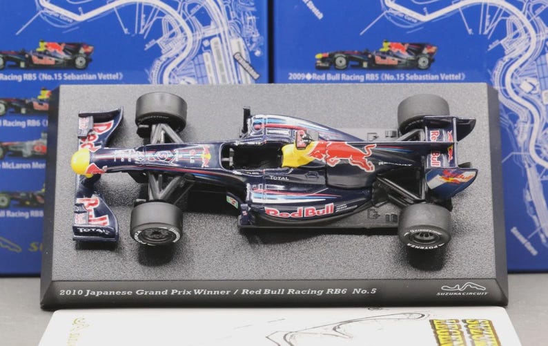 May include: A detailed model of a Red Bull Racing RB6 Formula 1 car. The navy blue car features red and yellow accents, sponsor logos, and the Red Bull logo. The model sits on a black base with text that reads "2010 Japanese Grand Prix Winner / Red Bull Racing RB6 No.5."
