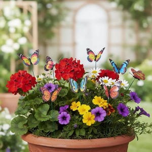 Puede incluir: Una maceta de terracota rebosante de flores vibrantes, incluyendo geranios rojos, margaritas blancas y petunias moradas. Estacas decorativas de mariposas en varios colores se insertan entre las flores, creando una escena de jardín caprichosa.