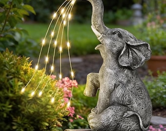 Puede incluir: Una estatua de jardín de elefante gris con la trompa levantada, que parece rociar un chorro de luces de hadas blanco cálido. La estatua está sobre una base de piedra, rodeada de vegetación y flores rosas, creando una pieza de decoración exterior caprichosa.