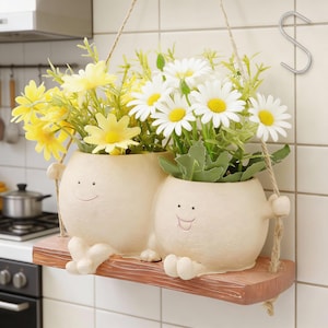 Może przedstawiać: Dwie kremowe donice w kształcie uśmiechniętych twarzy z żółtymi i białymi kwiatami, zawieszone na drewnianej półce za pomocą liny. Donice mają ramiona i nogi, tworząc kapryśny, dekoracyjny element do domu.