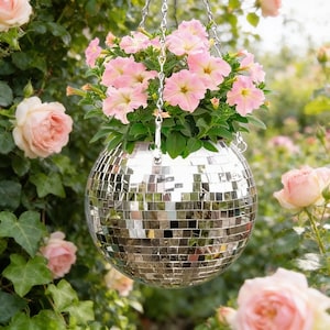 May include: A hanging disco ball planter filled with pink petunias. The spherical planter is made of mirrored tiles and suspended by a silver chain. The background features green foliage and soft pink roses, creating a whimsical garden scene.