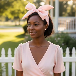 Puede incluir: Una mujer con una diadema rosa claro con un lazo grande y un vestido rosa a juego. El vestido tiene cuello en V y mangas cortas. El fondo incluye una valla blanca y vegetación.