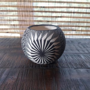 Puede incluir: Un cuenco de cerámica esférico con un patrón geométrico en blanco y negro. El diseño consiste en finas líneas radiantes que forman un motivo central de estrella, envolviendo la superficie exterior curva del recipiente.