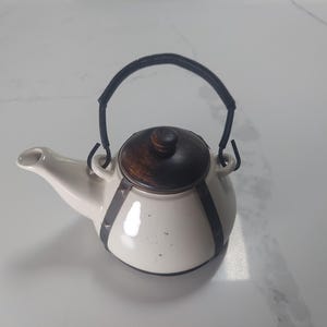 May include: A cream-colored ceramic teapot featuring a dark brown wooden lid and a black metal handle. The body of the teapot is accented with dark metal straps that extend from the base to the top, providing a structured appearance to the vessel.