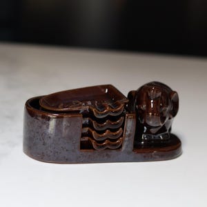 May include: A dark brown ceramic coaster set featuring a holder with a sculpted elephant figure on one side. The set includes four individual heart-shaped coasters stacked vertically within the holder, all finished with a glossy, speckled glaze.