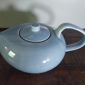 May include: A light blue ceramic teapot with a matching lid and curved handle. The teapot has a rounded body and a spout for pouring. The handle has a slight green hue. The teapot is designed for brewing tea.