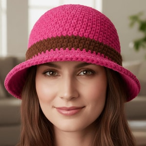 May include: A pink crochet cloche hat with a brown band. The hat has a rounded crown and a downward-sloping brim. The hat is made of a textured yarn and is a stylish accessory.