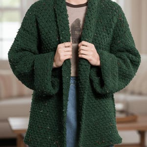 Könnte beinhalten: Dunkelgrüne, grob gestrickte Strickjacke mit weiten Ärmeln. Der offene Pullover hat ein strukturiertes Aussehen mit Farbtupfern. Die Strickjacke wird offen gehalten und zeigt ein T-Shirt und eine blaue Jeans.