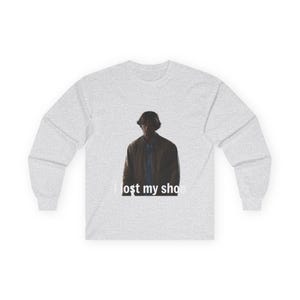 May include: A pale grey long-sleeved top featuring a graphic of a person in a brown jacket. The text "I lost my shoe" is printed beneath the image.