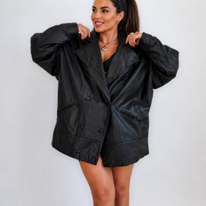 Puede incluir: Un blazer de cuero negro con diseño cruzado. La chaqueta tiene un cuello con solapas clásico, dos bolsillos delanteros y cierres de botones. El blazer parece ser de gran tamaño.