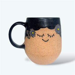 May include: A ceramic mug with a black speckled top and handle, and a tan speckled body. The mug features a smiling face with closed eyes and blue flower accents. The mug is designed for hot beverages.