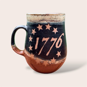 May include: A ceramic mug featuring a dark black and brown speckled glaze with a cream-colored rim. The front displays the text 1776 in a light tan color, encircled by a ring of twelve small, five-pointed stars in the same light tan hue.