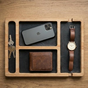 Può includere: Vassoio organizer in legno con scomparti contenenti uno smartphone, chiavi, un portafoglio in pelle marrone e un orologio da polso con cinturino in pelle marrone. Il vassoio ha una finitura in legno chiaro e scomparti foderati in feltro nero.
