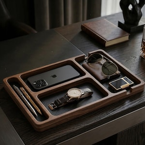 以下が含まれることがあります： 複数のコンパートメントを備えた木製のデスクオーガナイザー。スマートフォン、サングラス、財布、時計、ライター、ペンが入っています。オーガナイザーは濃い色の木材で作られ、黒い革のようなベースが付いています。