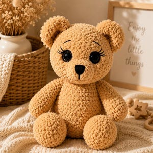 May include: A handmade, tan crochet teddy bear with large black eyes and long eyelashes. The bear has a black nose and is sitting on a cream-colored knitted blanket. The bear is a soft toy.