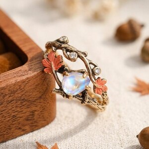 Peut inclure: Une bague dorée avec un design inspiré de la nature. La bague présente une pierre précieuse irisée en forme de poire, ornée de petites pierres claires et de feuilles rouges stylisées. La bande a une texture de branche. Des glands et des feuilles sont dispersés autour de la bague.