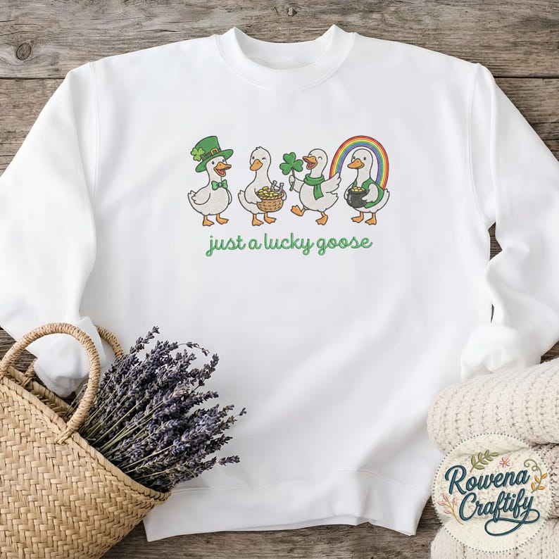 May include: White sweatshirt with a St. Patrick's Day theme. Features a graphic of four cartoon geese, each holding a St. Patrick's Day item, with a rainbow and the text "just a lucky goose" in green.