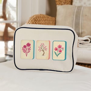 Puede incluir: Un estuche rectangular de lona color blanco hueso con ribetes azul marino oscuro en los bordes. La parte frontal presenta tres azulejos rectangulares bordados, cada uno con un dise&ntilde;o floral rosa diferente: un rosal, un cerezo y una orqu&iacute;dea.