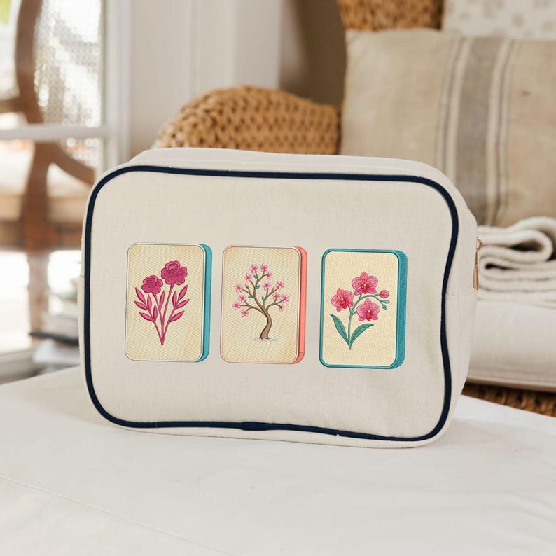 Puede incluir: Un estuche rectangular de lona color blanco hueso con ribetes azul marino oscuro en los bordes. La parte frontal presenta tres azulejos rectangulares bordados, cada uno con un dise&ntilde;o floral rosa diferente: un rosal, un cerezo y una orqu&iacute;dea.