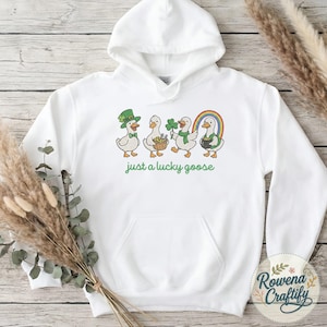 May include: White hooded sweatshirt with a St. Patrick's Day theme. The design features four cartoon geese, each with festive accessories like a leprechaun hat, basket of gold, shamrock, and rainbow. The text "just a lucky goose" is printed below the geese.