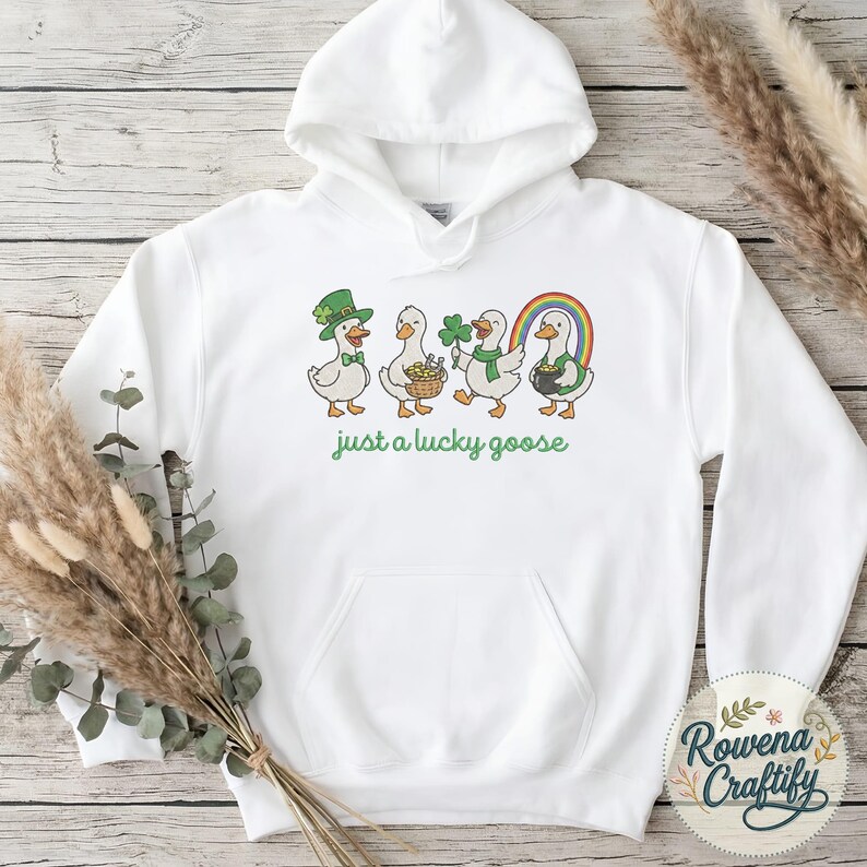 May include: White hooded sweatshirt with a St. Patrick's Day theme. The design features four cartoon geese, each with festive accessories like a leprechaun hat, basket of gold, shamrock, and rainbow. The text "just a lucky goose" is printed below the geese.